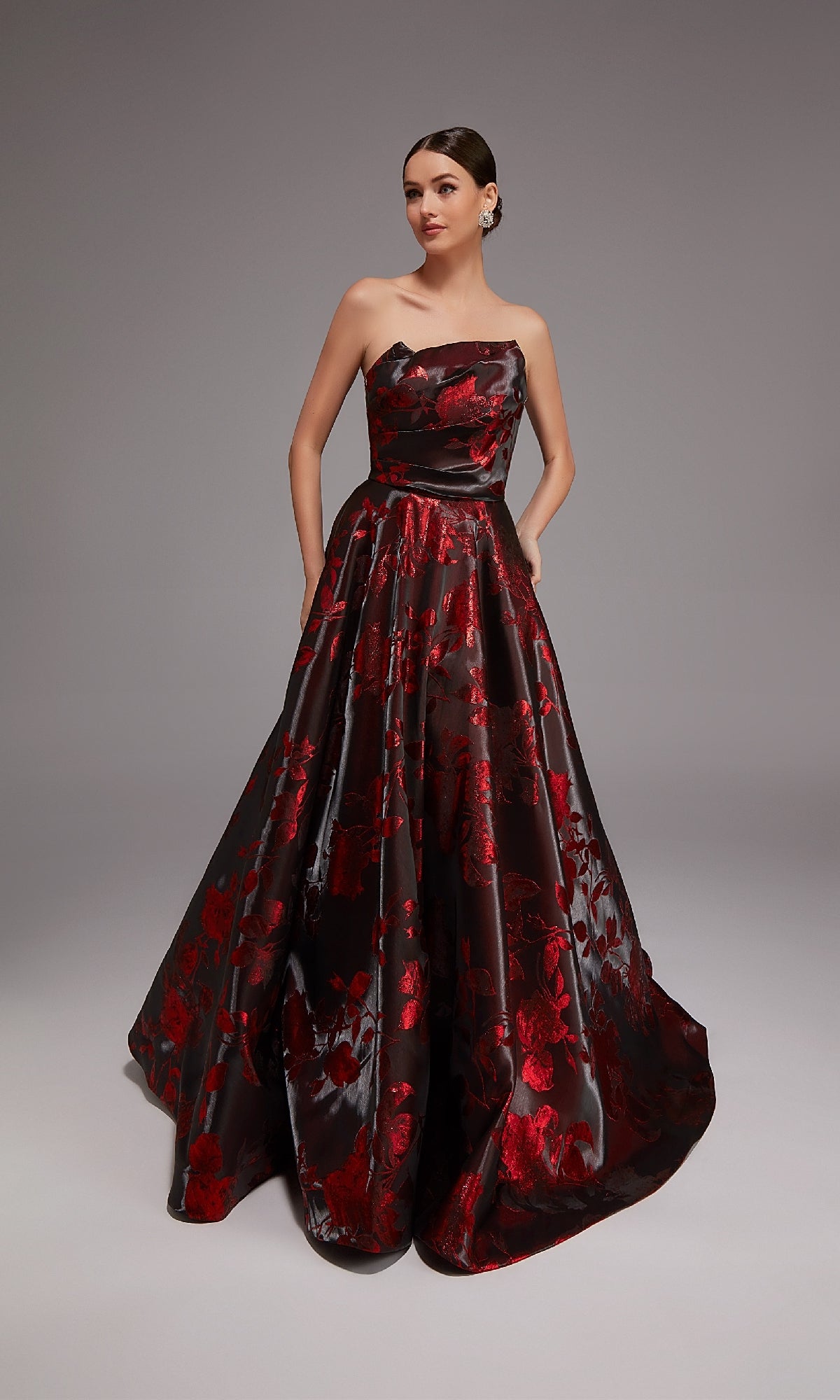 A woman stands against a plain gray background wearing the Alyce 27862 long formal strapless black jacquard dress with a red floral pattern.